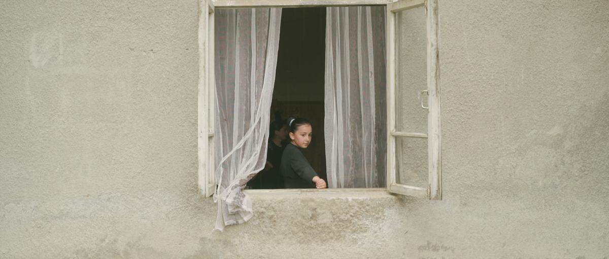 A young girl stares out the open window of a rustic stone building