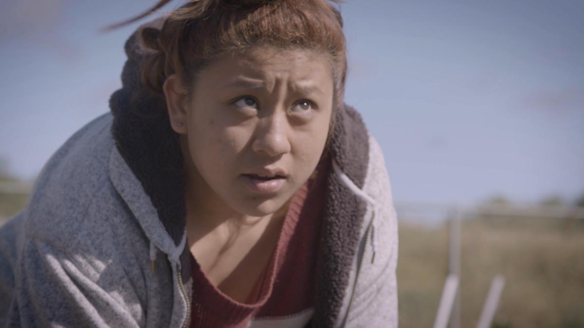 A Latina teenager leans over in a field, tilting her head upwards and looking away from her harvesting work.