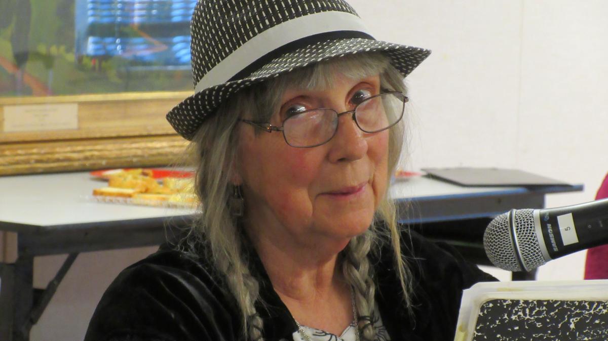 An older White woman in glasses and hat reads from a composition book before a notebook.