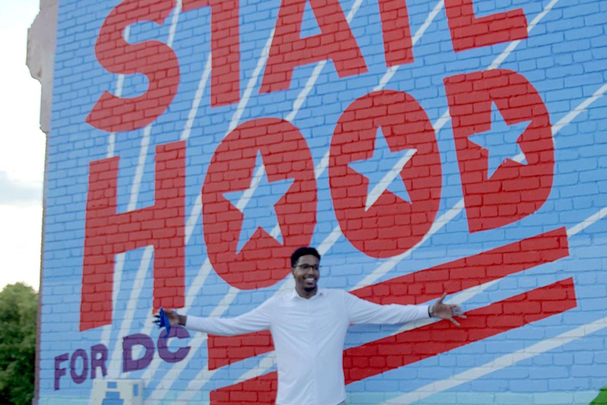 A young Black man smiles with his arms spread wide in front of a blue and white mural reading "statehood."