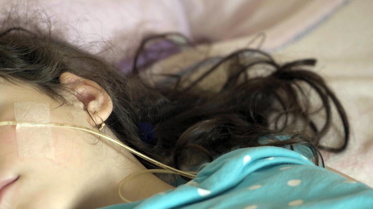 A girl with dark brown hair is lying on the bed, being nourished via feeding tube