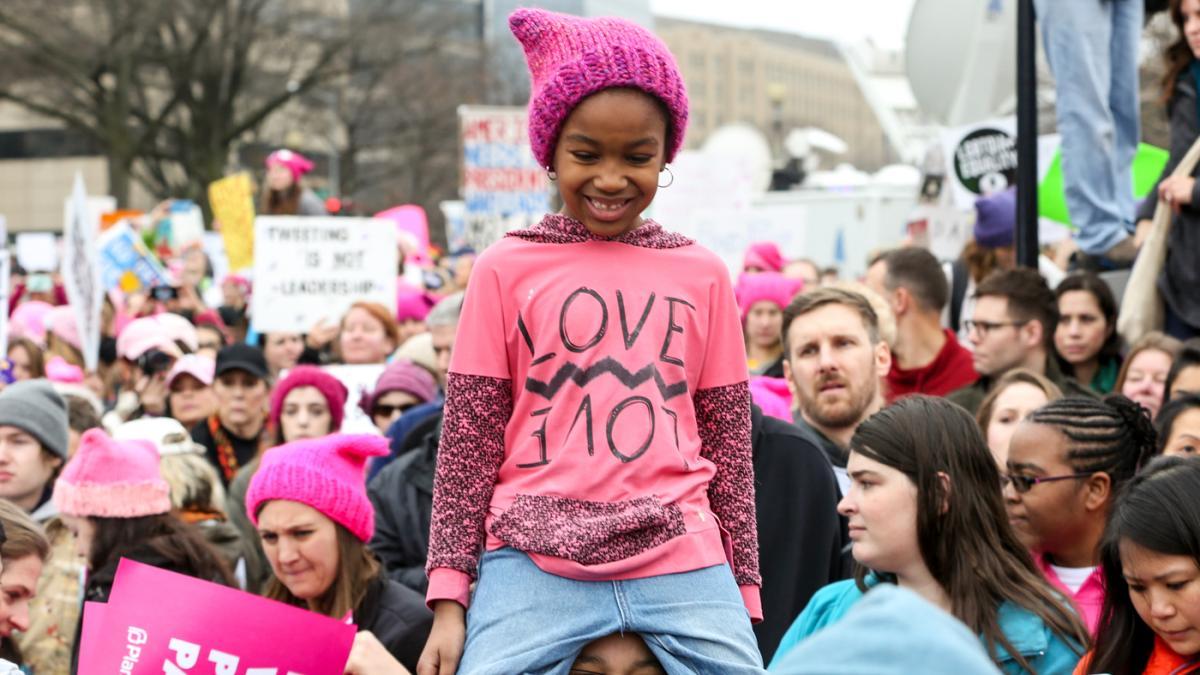 A feminine african-american child in a pink "pussyhat" on someone's shoulders during a woman's rights march. 