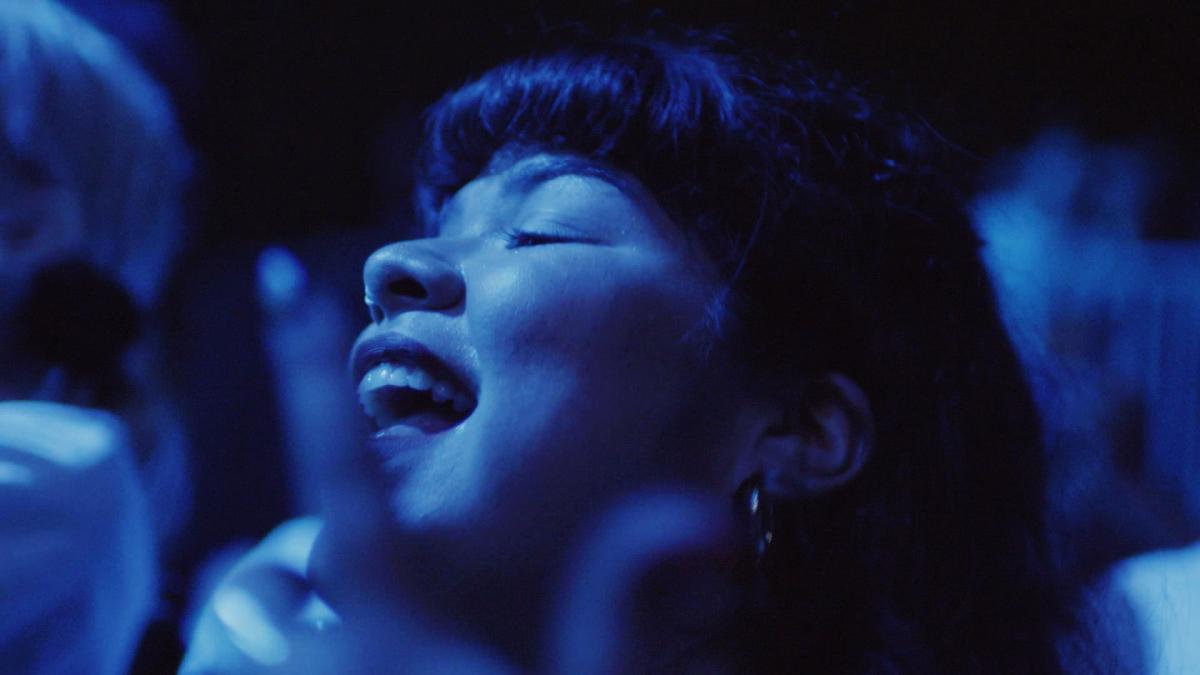 Latina woman dances to music amongst a crowded concert audience, in blue light. 