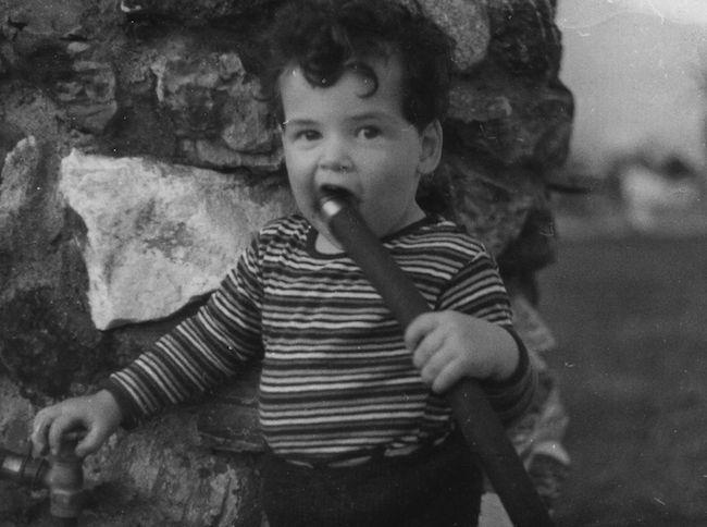 A white male toddler stands in front of a stone wall, chewing on a garden hose.