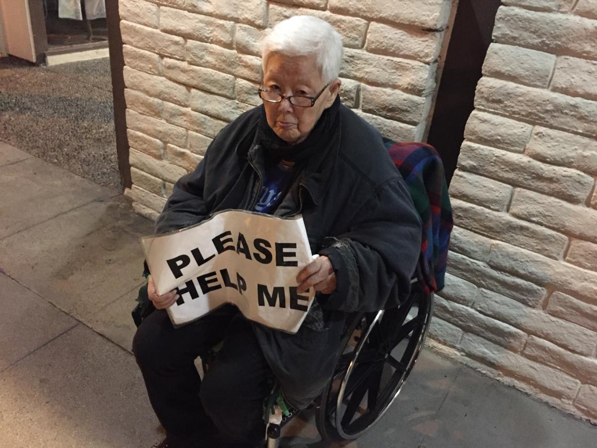 an elderly asian individual in a wheelchair holds a sign reading "please help me"