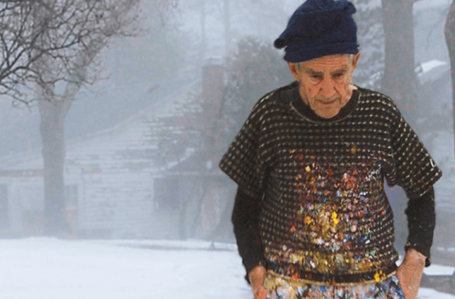 an older White Man with a paint-splattered sweater and knit cap stands in front of a snow-covered house.