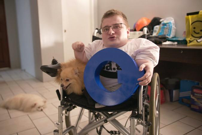 Fredrick Brennan sitting in wheelchair with his dog holding a blue 'Q' sign