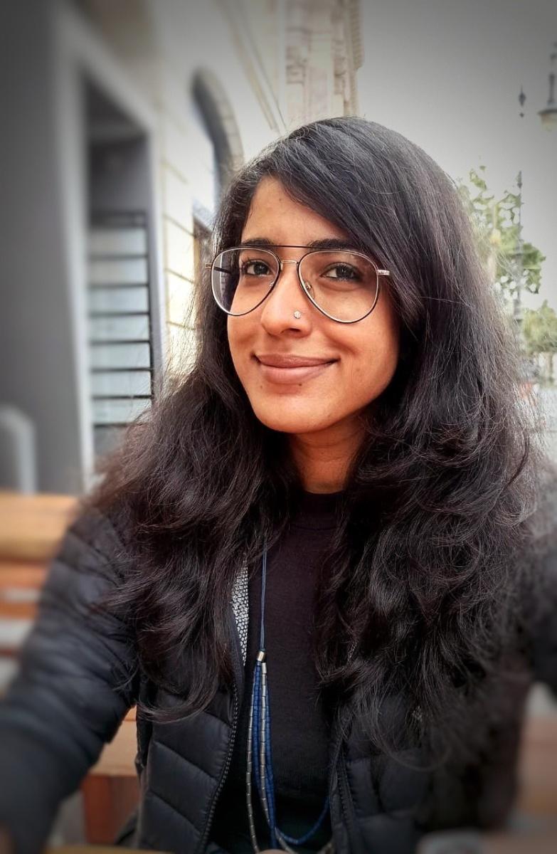 Woman of Indian descent with long black hair and glasses takes a selfie with a blurred background