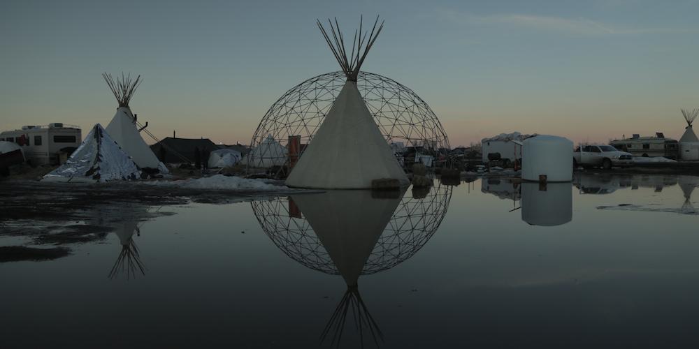 a white tipi sits in front of a steel geometric dome, both are reflected in a shallow pool at sunset.
