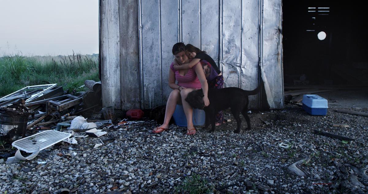 two female siblings sit on a plastic box and pet their black dog in front of a rural dilapidated barn.