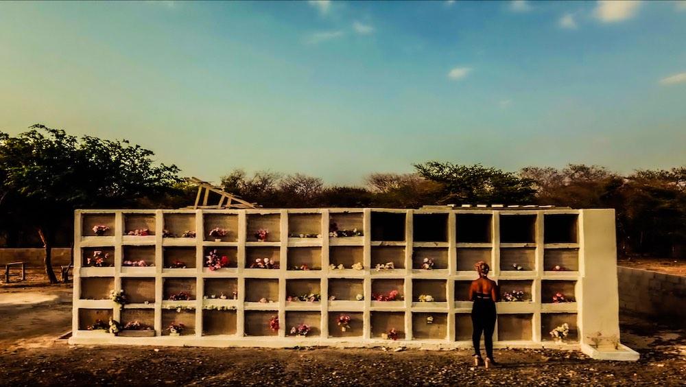 Sonia, a woman with dark skin, stands in her cemetery. She looks at a wall of graves. The text "People Like Us" is overlaid on top.