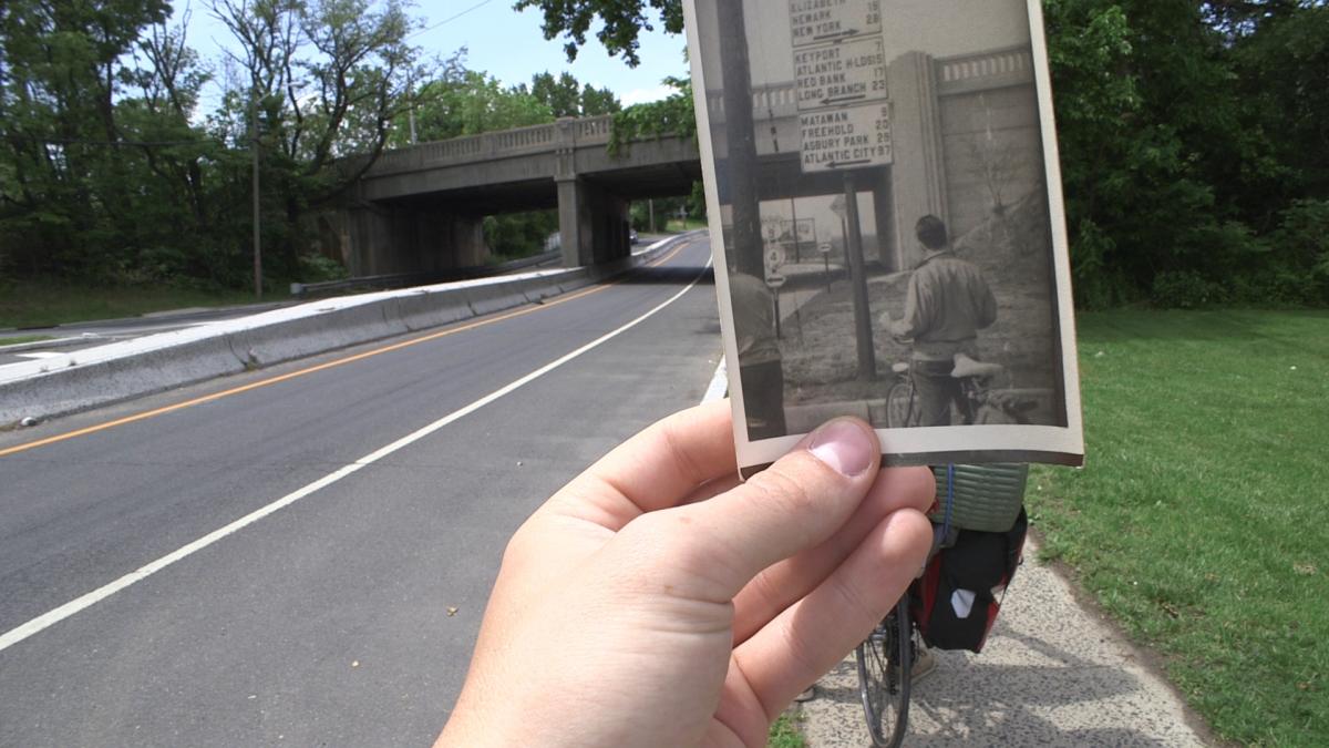 A hand holds an old photograph of a man with a bicycle at South Amboy Bridge. The photo lines up perfectly with South Amboy Bridge in the background. 
