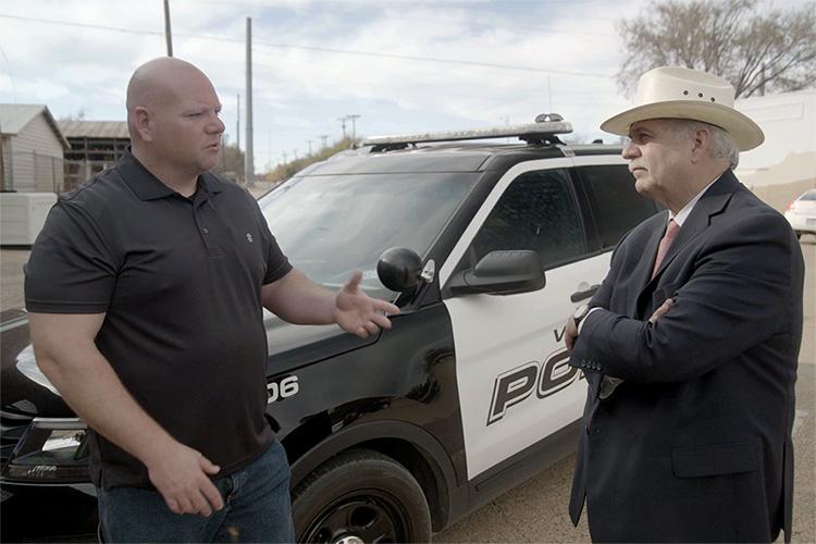A man in a black shirt and a man in a suit and hat talk in front of a police cruiser.