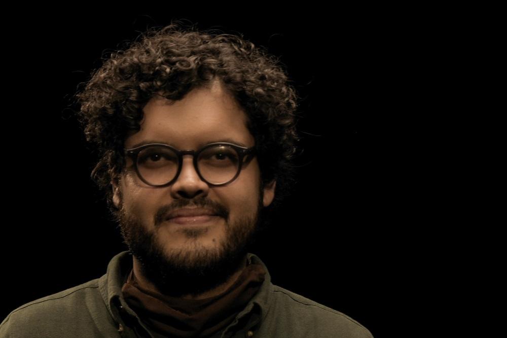 Manuel Acuna is pictured before a black background. He has curly dark brown hair, mustache and a beard. He is wearing black framed glasses and an olive green casual shirt.