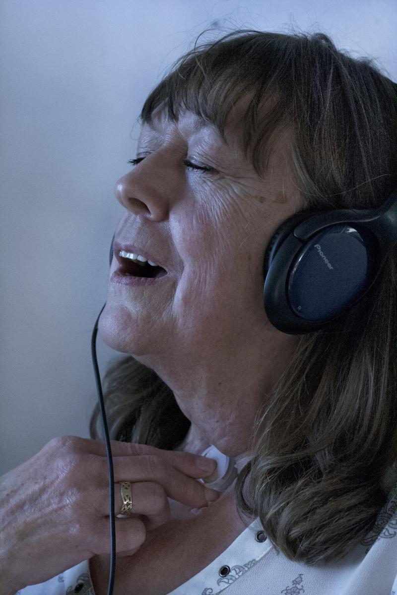 A middle-aged white woman with blue headphones closes her eyes as she adjusts a voicebox on her neck.