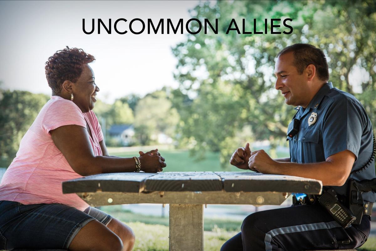 A Black woman sits across a park picnic table from a White Cop in uniform