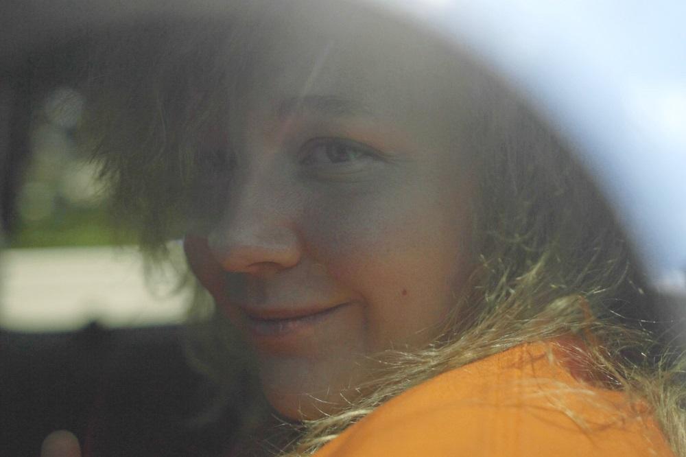 A still from "United States vs. Reality Winner": A young woman with blonde hair and an orange sweatshirt looks out from the window of a car.