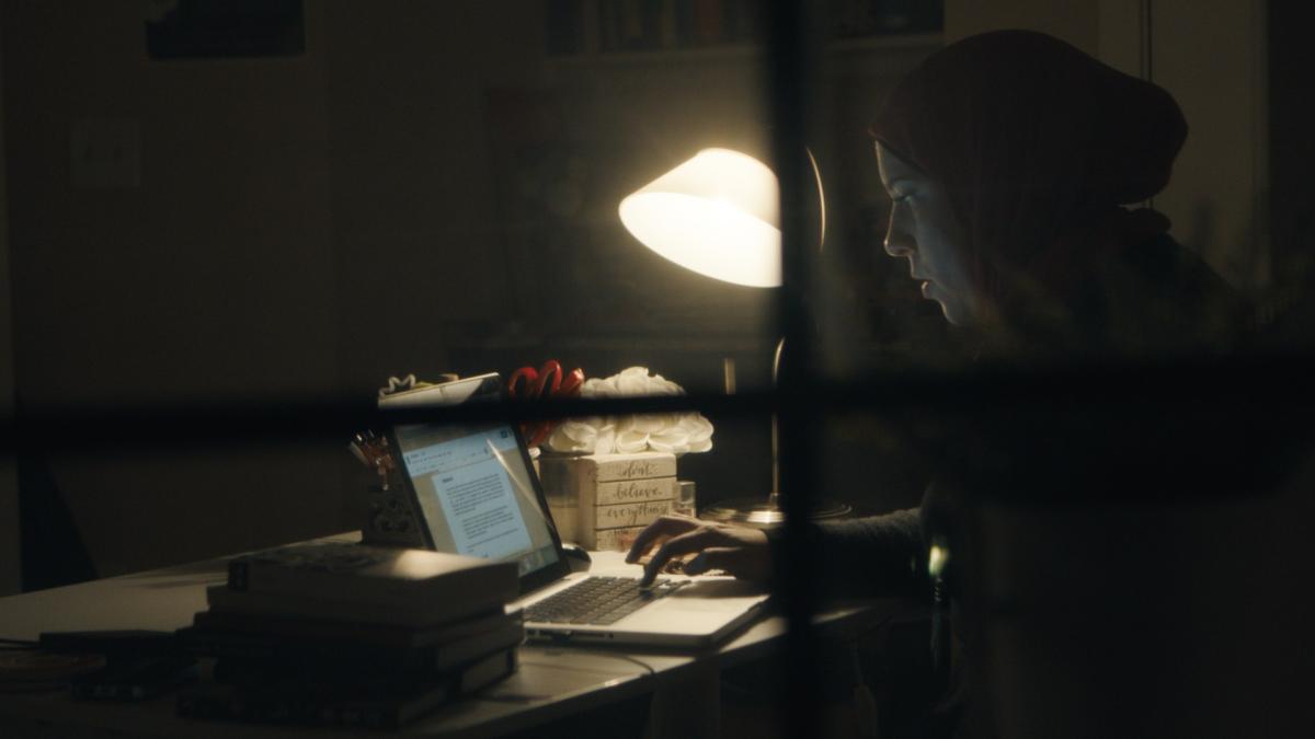 A woman in a hijab working at her computer late into the night, slightly obscured by a windowpane in the foreground. 