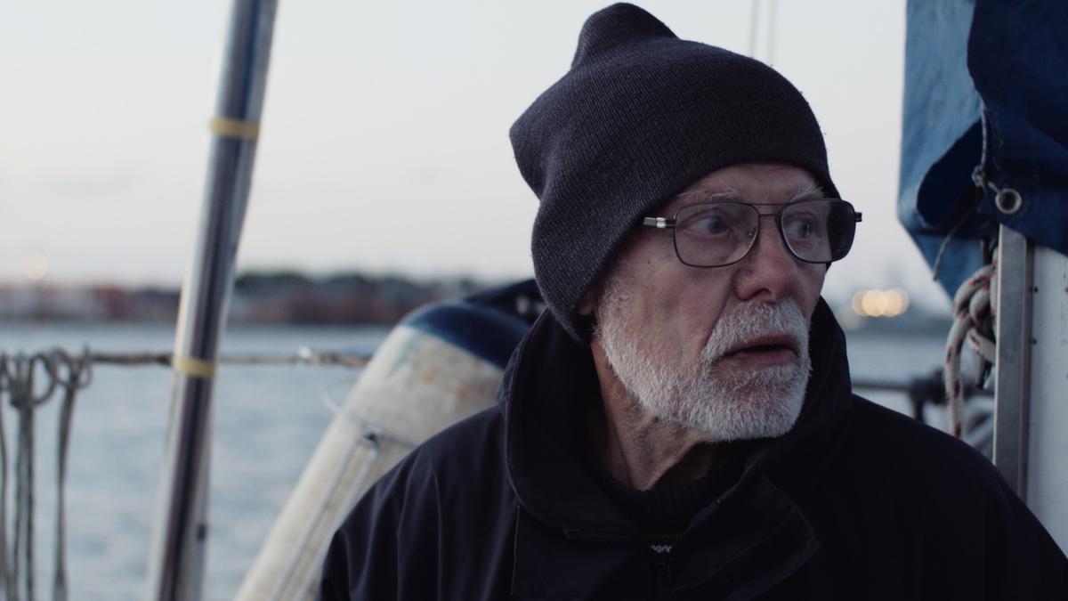 an older White man in glasses and a knit cap stands on the deck of a sailboat