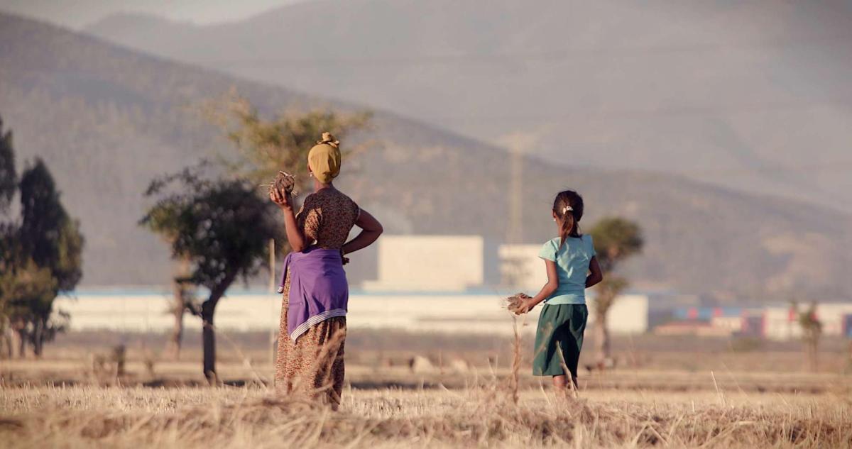 An Ethiopian farmer wearing a yellow headscarf and flowery dress and her daughter in ponytail wearing a blue T-shirt and green skirt stand in dry teff field looking into the distance against a hazy background of a massive industrial zone. 