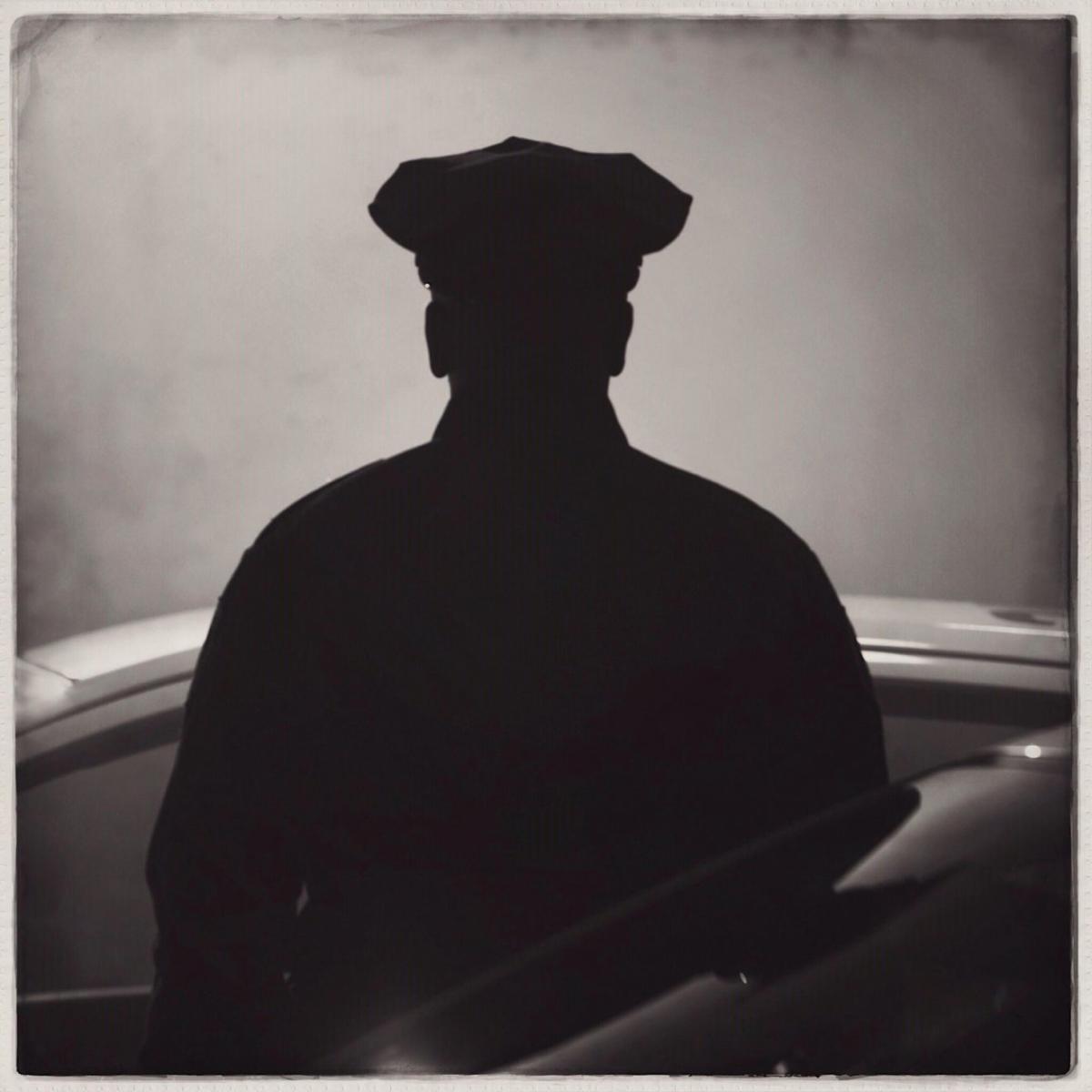 the black silhouette of a police officer stands between two parked cars. 