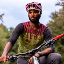 A Black man wearing a helmet sits on a bike. There are trees in the background.