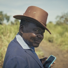 Photo courtesy of CPH:DOX. Still from 'Theatre of Violence.' A person with dark skin, wearing a brown rimmed hat and a blue coat, holding a cellphone, stands in a green field with a dirt path.
