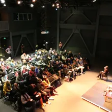 The wide angle image shows an group of more than 100 people are sitting facing Stanley Nelson who has a microphone in hand is presenting with light spotlighting him.