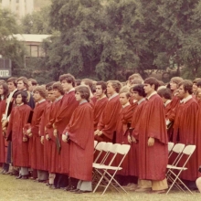 A group of several dozen young men stand on a field wearing red graduation robes. They are holding diplomas.  