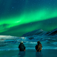 Two people, surely avid adventurers, stand waist deep in a body of arctic water; maybe they’re in Alaska - gazing upon a vast night sky. Most notably there is a bright, luminescent green aurora borealis shining above them. 