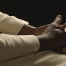This picture shows a close-up of dark-skinned hands belonging to a Catholic nun dressed in a white robe against a black background. Her face can not be seen. The area in which the shot was taken is an interior illuminated with artificial light creating a high contrast.