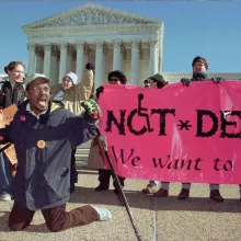 A Black man leads a group of protesters in front of the Supreme Court, kneeling with his crutches outspread and his mouth mid-yell, while holding a small American flag. A neon pink banner to the right reads, "NOT DEAD. We want to live!"
