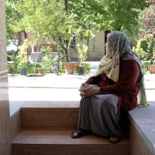 A female person, the main protagonist of the film, Hawa, sitting on the stairs of he appartement. She is looking away from the camera, watching a green garden. Hawa is wearing a colourful scarf covering her head, a red jacket and a grey dress.