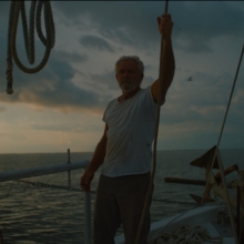 A nearly silhouetted silver-haired man stands on the bow of a boat, holding onto a rope above his head. Behind him is a cloudy dusk sky and an expanse of water.