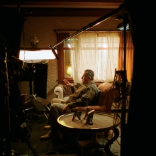 A wide angle shot of a Latino man seated in a living room surrounded by documentary interview equipment.
