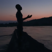 Film still from 'Eat Bitter,' showing the silhouette of a man with arms outstretched, on a small boat.