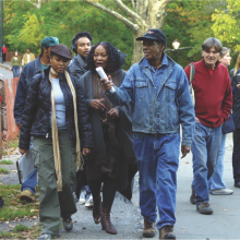 Greaves wearing a black hat with a blue jean jacket and blue jeans, with cast and crew in Central Park during shooting of Symbiopsychotaxiplasm: Take 2 1⁄2 (2003).