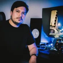 A man sitting in front of a color grading suite looks at the camera. He has short black hair and brown eyes and is wearing a black T-shirt.