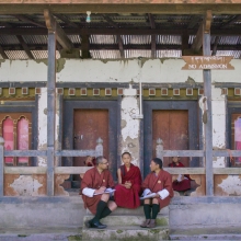 Still image from 'Agent of Happiness,' depicting three male figures seated on the steps of an ornate temple. All three are wearing robes in a deep maroon.