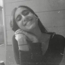 Black and White image of a woman with brown skin smiling at the camera with her head tilted. A woman with white skin smiles at the camera. She has mid-length, straight black hair and is wearing a black top.