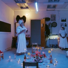 Still of a group of people in white dresses, conducting a ritual in a darkened studio.