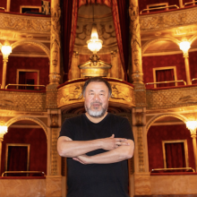 An Asian man wearing a black shirt stands with his arms crossed in front of the balcony seating of an opera house