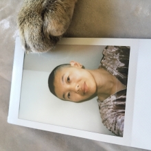 A photo of an androgynous person of Asian descent with a light smile at the camera. They have a dark shaved head and brown eyes and are wearing a pink velvet v-neck. The photo frames a photo print from a Polaroid camera; the print is lying flat on a silver texture, with a cat's paw gently lying on the top right corner of the print.