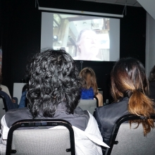 Photograph of the back of an audience, watching a screen hung on a black wall in front of them.