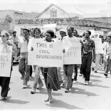 Black and white image of a group of people marching and holding protest signs. One sign says, "THIS IS CIVIL DISOBEDIENCE."