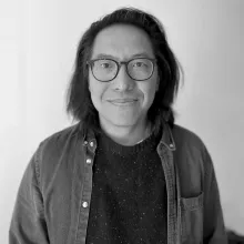 Black and White image of an Asian man. He is wearing black glasses and smiles at the camera. He has shoulder-length black hair and is wearing a dark sweater and button-down shirt.