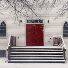 Snowy stairs lead up to a symmetrically-centered bright, red church door with a frosted church window on each side, and bare, snowy branches reaching toward the door from above