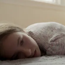 Close up of a teenage girl with light-brown hair and blue eyes, wearing a light-pink sweater and jeans, laying on her stomach on a bedroom floor with a noticeable, hunched-over spine, looking melancholily away from the camera.