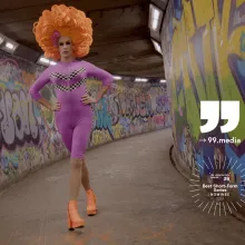 A colourful drag queen, wearing an orange foam wig, a purple leotard and high heels, marches through a Belfast tunnel whose walls have been spray-painted.