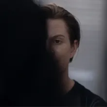 The image depicts two people facing one another. The back of one woman's head and her long black hair fill the left half of the frame. The black hair obscures half of another woman's face who is facing the camera.